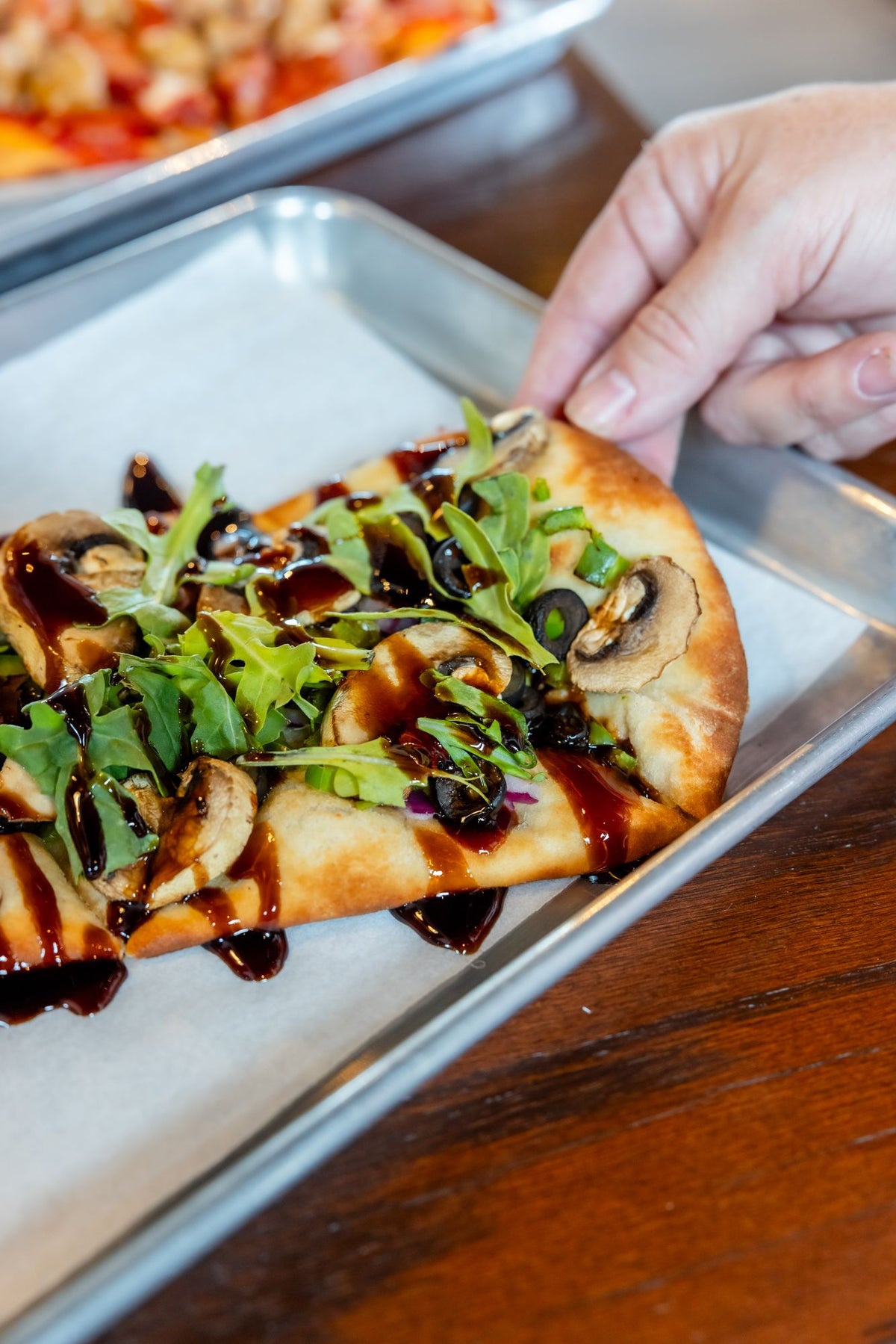 A flatbread pizza with arugula, mushrooms, olives, and a balsamic drizzle, lifted from its tray