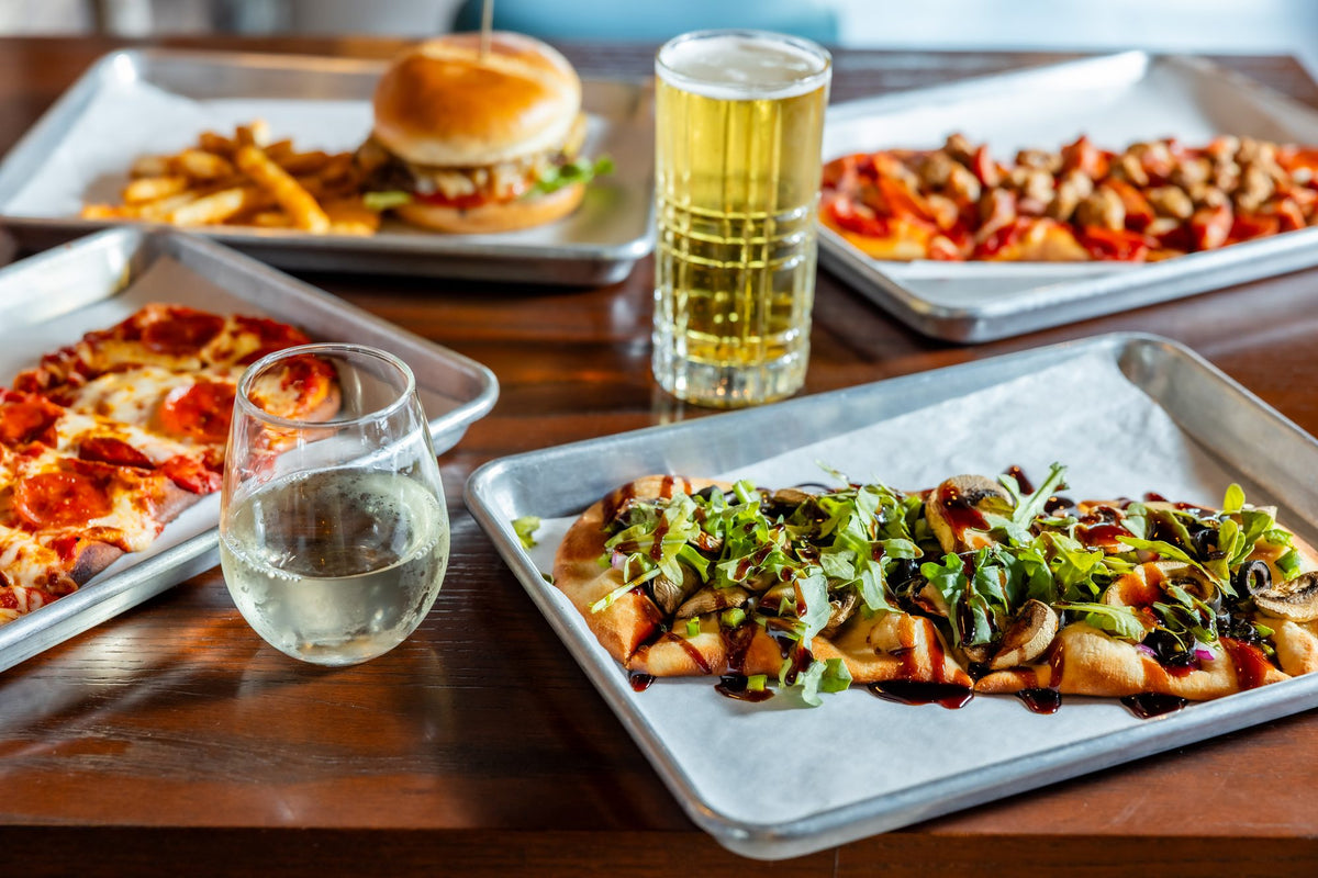 A burger and fries, flatbread pizzas, a beer, and a glass of white wine on a wood table at The Mantel