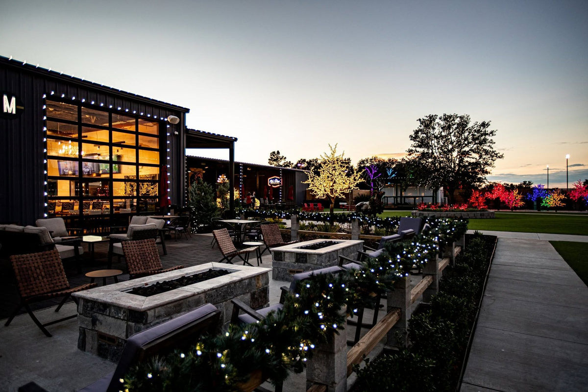 The patio at dusk, with stone fire pits, garland lights, and lit trees beyond